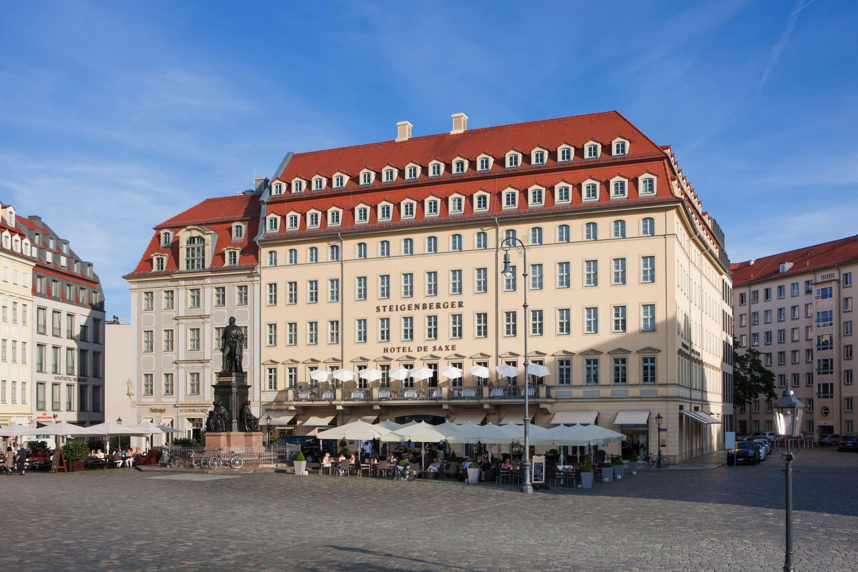 Steigenberger Hotel De Saxe Dresde Exterior foto The photo shows a large, elegant building with a light-colored facade and a distinct red roof. The structure features multiple windows and a decorative style typical of historic architecture. In front of the building, there is a plaza with outdoor se